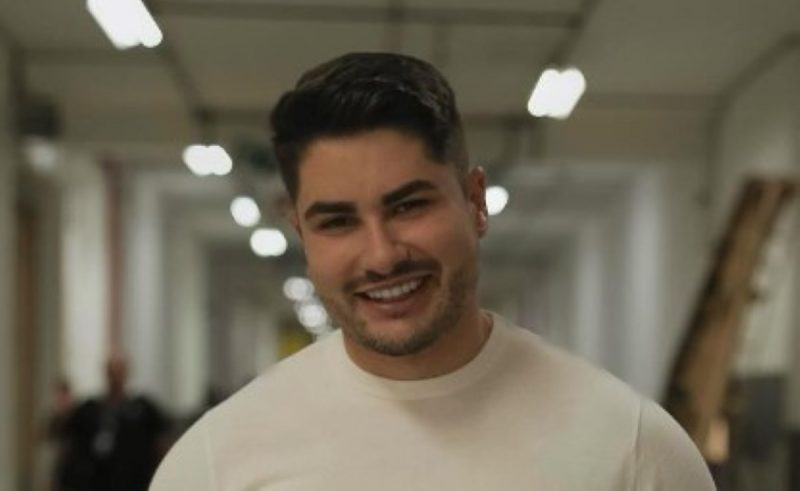 Na foto Lucas Souza aparece sorrindo com uma camiseta branca e olhando para frente 