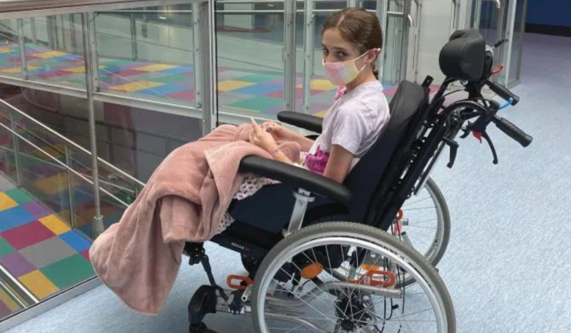 Foto mostra menina sentada em cadeira de rodas enquanto usa máscara no rosto