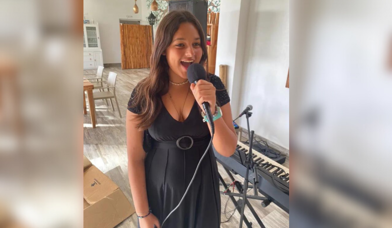 Foto mostra menina com a pele parda, cabelos escuros e longos na altura dos seios, sorrindo enquanto canta no microfone. Ela usa vestido preto com decote e está de pé ao lado de teclado