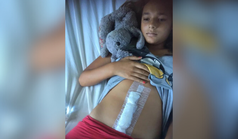 Foto mostra menina com expressão triste deitada em cama com lençol branco. Ela segura a mão na blusa e levanta para mostrar curativo de cicatriz na barriga.