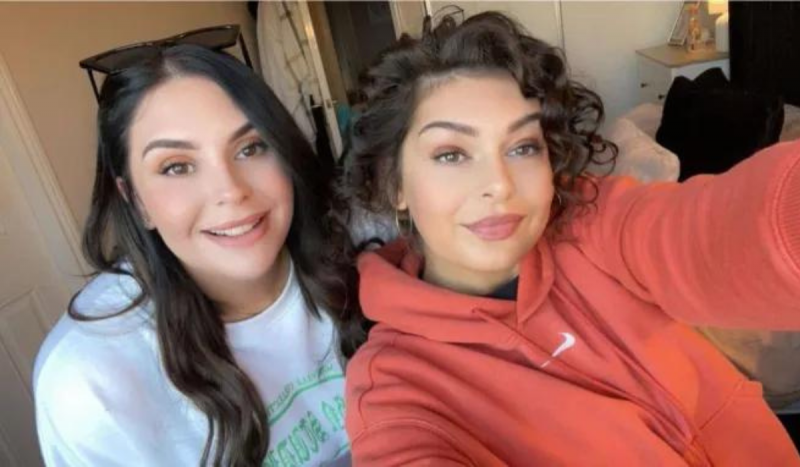 Duas mulheres brancas de cabelos escuros posam para foto fazendo selfie. Mulher à esquerda da foto usa camisa branca e óculos escuros na cabeça e mulher à direita usa moletom na cor coral