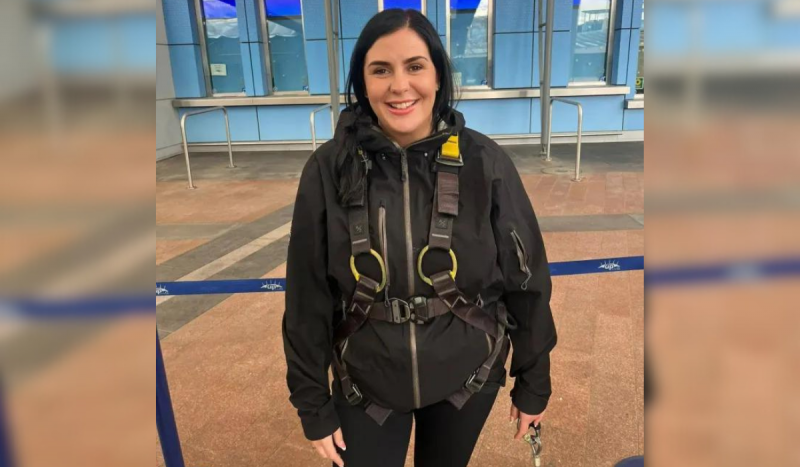 Mulher branca sorri de pé enquanto usa roupa preta com o que parece ser equipamento de segurança por cima