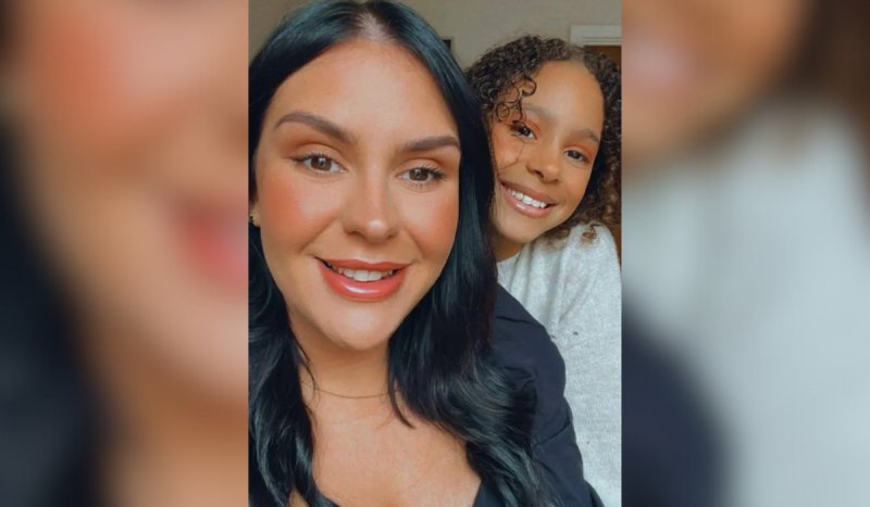 Mulher branca de cabelos pretos sorri. Ela está ao lado de uma criança negra de cabelos cacheados que também está sorrindo