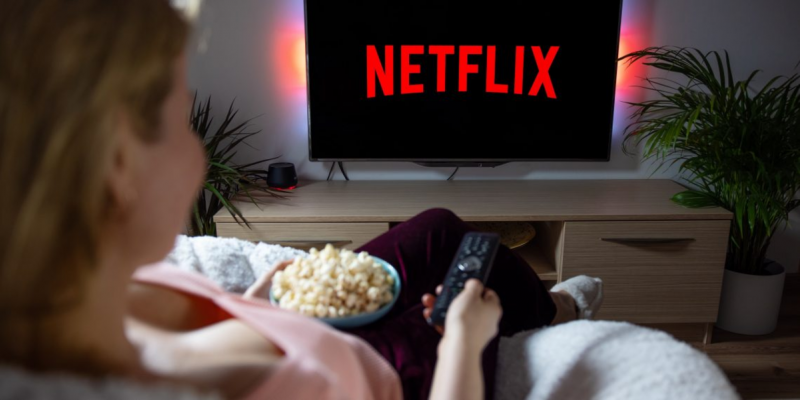 Mulher vendo Netflix com pipoca e controle na mão para matéria sobre truque para ter acesso a mais filmes e séries na Netflix.