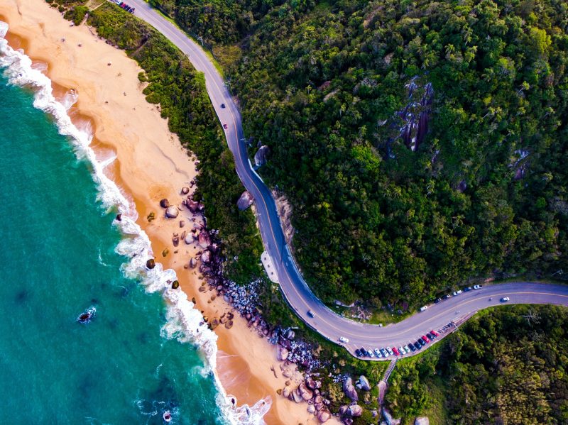 Foto aérea de um trecho da Rodovia Interpraias em Balneário Camboriú. Bem no centro da foto, a via de asfalto e uma de suas curvas, onde alguns carros estão parados no acostamento. Do lado direito, um vasto morro coberto por vegetação. Do lado esquerdo, aparece parte do mar de águas esverdeadas e a faixa de areia.