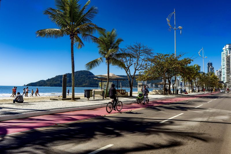 Foto da orla da Praia Central de Balneário Camboriú. A foto mostra parte da via de carros, a ciclofaixa pintada de vermelho, onde andam dois ciclistas e um homem caminha, a calçada e a praia. Pela orla, várias árvores plantadas. Ao fundo, o mar e alguns morros verdes. 