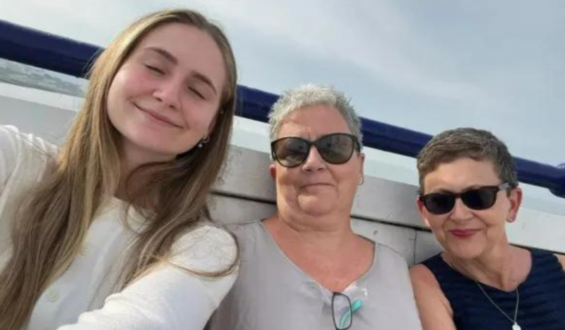 Foto mostra três mulheres. À esquerda jovem loira de cabelos compridos sorrindo com lábios e olhos fechados de blusa de mangas branca; ao centro mulher branca com expressão séria olhando para frente usando óculos escuros; e à direita mulher branca com óculos escuros e blusa azul escuro