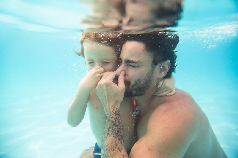 Foto de um homem jovem, de pele clara, cabelos e barba preta, ao lado de uma criança de pele clara e cabelo loiro. Eles estão abraçados e submersos na água de uma piscina do parque aquático Cascanéia. Estão de olhos fechados e tapam o nariz. 