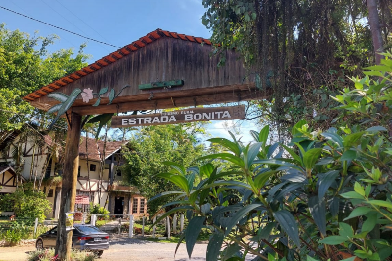 Foto do portal de entrada da Estrada Bonita. O portal é feito com dois troncos de madeira e um pequeno telhado em duas águas. Há uma placa também em madeira onde está escrito em branco: Turismo Rural Estrada Bonita. No canto direito há folhagens e vegetação. Do lado esquerdo aparece parte da estrutura de uma pousada. 