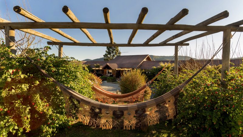 Foto de uma das pousadas em Urubici, a pousada Fogo Eterno. Em primeiro plano, aparece um gazebo feito de troncos de madeira, espaçados entre si e onde estão penduradas duas redes em tons de bege e marrom. Ao redor delas, há arbustos. Ao fundo, aparece parte do grande prédio de entrada da hospedagem. 