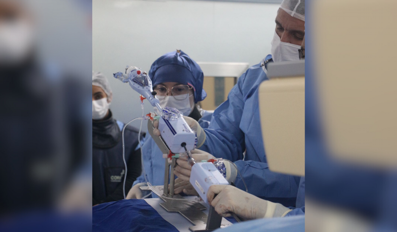 Profissionais da saúde em centro cirúrgico fazendo cirurgia no coração de paciente. Eles usam roupa e toucas azuis