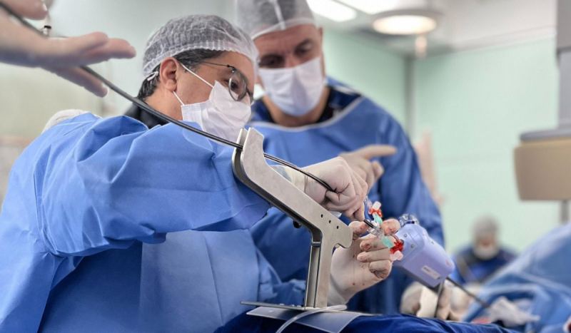Médicos segurando objeto cirúrgico dentro de sala hospitalar. Eles usam roupa azul e toucas descartáveis de cor branco. Foto é usada para ilustrar procedimento com técnica menos invasiva em Florianópolis