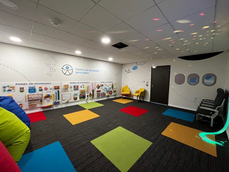 Foto da sala multissensorial do Aeroporto de Florianópolis. O chão é de carpete com quadrados coloridos. No canto esquerdo, pufs coloridos, nas cores vermelho, verde e azul. Em uma das paredes há um painel de atividades interativo. 