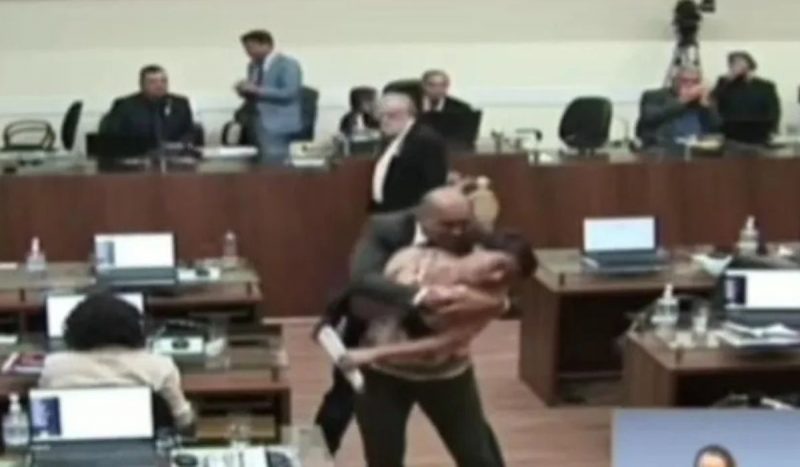 vereador Marquinhos aparece abraçando e vereadora Carla Ayres por trás no meio da Câmara de vereadores, enquanto ela tenta se afastar