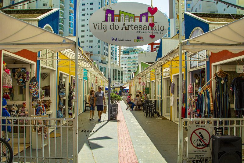 Foto da Vila do Artesanato, no Centro de Balneário Camboriú. Na parte superior e centralizada, uma placa branca onde está escrito Vila do Artesanato e desenho de casas coloridas. Abaixo dela, há um portal branco aberto. Atrás estão duas fileiras de pequenas lojas coloridas com alguns artigos expostos do lado de fora. Entre as lojas, há uma calçada. Um casal anda de mãos dadas e um grupo de pessoas está sentado em uma mesinha. Ao fundo, aparecem alguns prédios. 