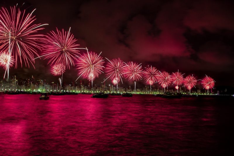 Foto do show de fogos na Praia Central de Balneário Camboriú. Na foto, aparecem 10 grandes círculos luminosos de cores vermelha e branca no céu. Atrás deles aparecem as luzes da orla da Praia Central e prédios iluminados. À frente, está o mar que reflete a cor vermelha dos fogos. 