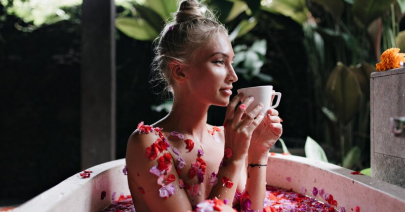 Mulher loira tomando liquido em xícara em banheira com várias pétalas de flores em seu corpo