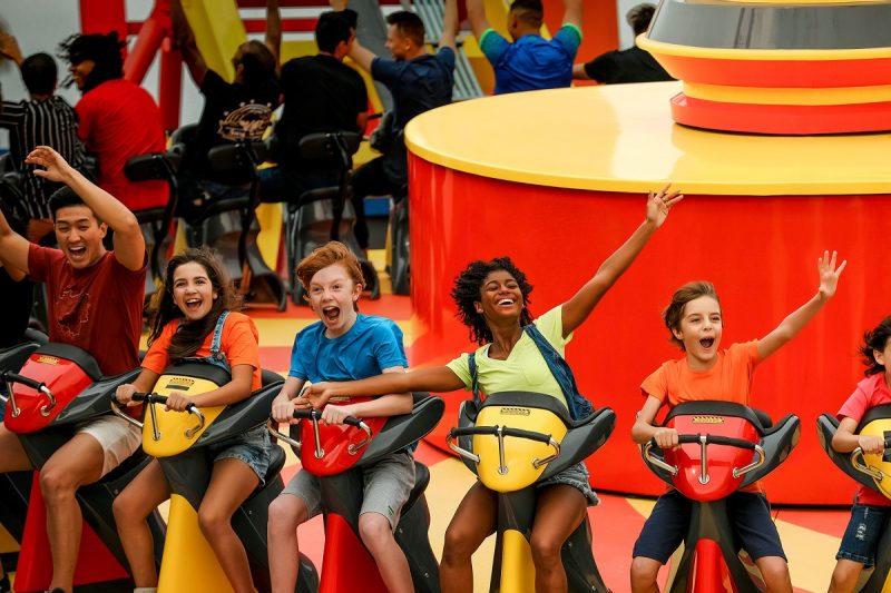 Três homens jovens e duas mulheres jovens, de diferentes idades e características físicas, estão sentados em um grande disco giratório laranja e amarelo no Beto Carrero World. Todos sorriem e três deles estão com os braços erguidos. 