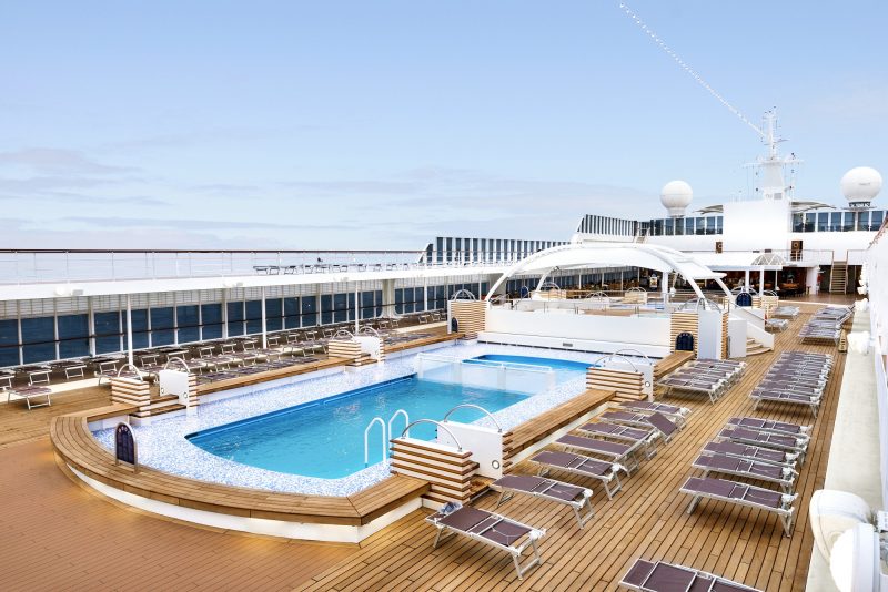 Foto de um amplo deck de madeira com uma grande piscina no centro em um navio. Ao redor há várias espreguiçadeiras. O céu está azul. 