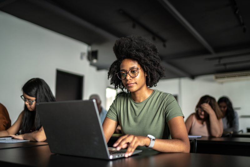 Mulher de camisa verde, sentada em frente a uma mesa com notebook. Ao fundo, outras pessoas estudando.