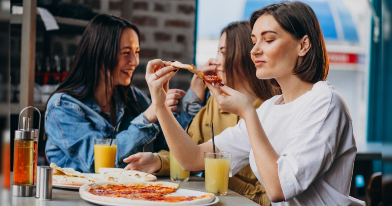 Três mulheres, uma de jaqueta jeans, blusa amarela e outra de blusa branca, comendo pizza no Dia da Pizza