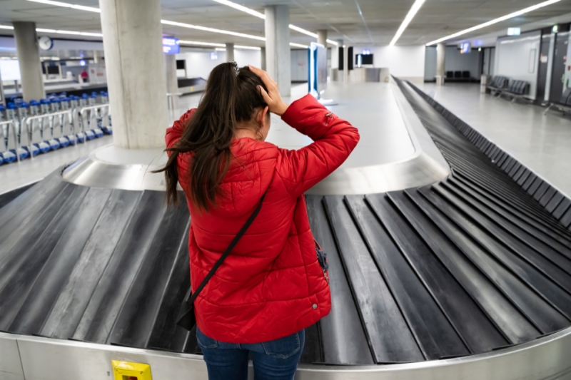 Foto interna da área de desembarque de um aeroporto. No centro da foto, uma esteira de bagagens, sem nenhuma mala. Uma mulher jovem, de pele clara e cabelo preto comprido está com as mãos na cabeça enquanto olha para a esteira. Ela usa uma japona vermelha e calça jeans e está de costas para a foto. 