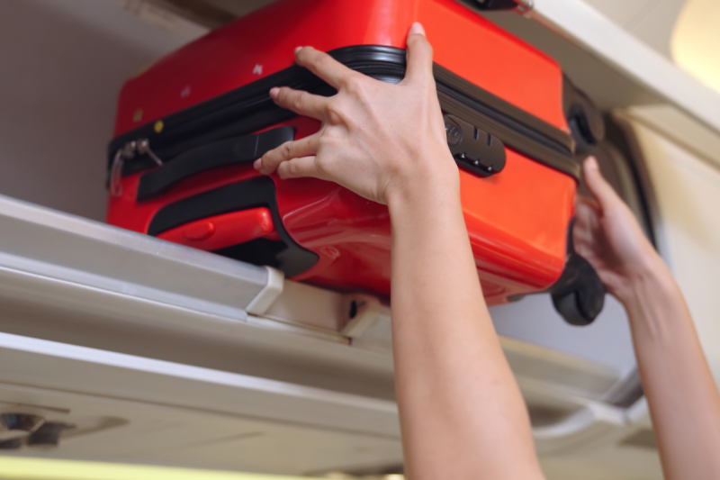 Foto das mãos de uma mulher jovem e de pele clara colocando no bagageiro da cabine do avião uma pequena mala vermelha. 