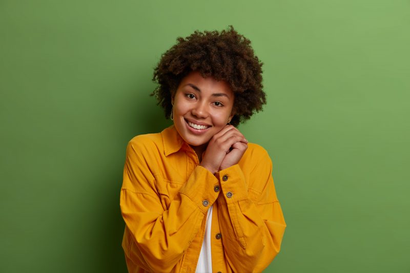 mulher negra sorrindo, ela está com uma blusa amarela e as mãos unidas ao lado do rosto com um sorriso nos lábios representando as oportunidades da Lua em Escorpião