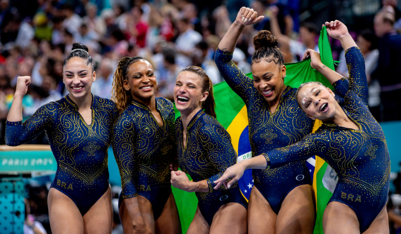 Julia Soares, Rebeca Andrade, Jade Barbosa, Lorrane dos Santos e Flávia Saraiva, respectivamente, a equipe da ginástica brasileira feminina - foto mostra cinco mulheres vestindo collant azul-marinho com brilhos dourados sorrindo e comemorando vitória