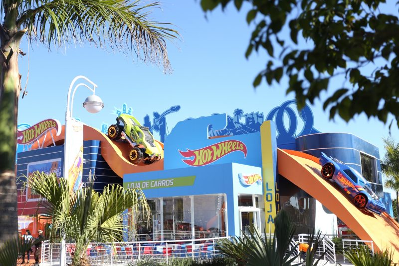 Fachada da área temática da Hot Wheels no Beto Carrero World. A fachada é azul, com o nome escrito em amarelo e dentro de uma faixa vermelha em formato de chama de fogo. Por cima da fachada há uma pista laranja com dois carros da Hot Wheels. A fachada aparece por entre folhagens de árvores. 