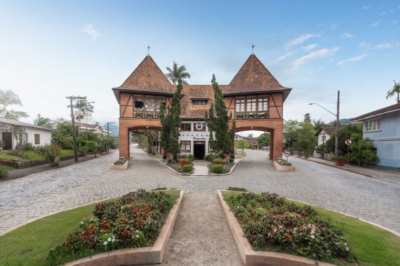 Foto do Portal Sul da cidade de Pomerode. O portal é em estilo germânico, construído em tijolos à vista, com ripas de madeira cruzadas, como em construções enxaimel, e telhado pontiagudo. Na parte de cima há janelas de madeira. Em baixo há um vão por onde passam carros. Na frente do portal há um canteiro com flores e arbustos. 