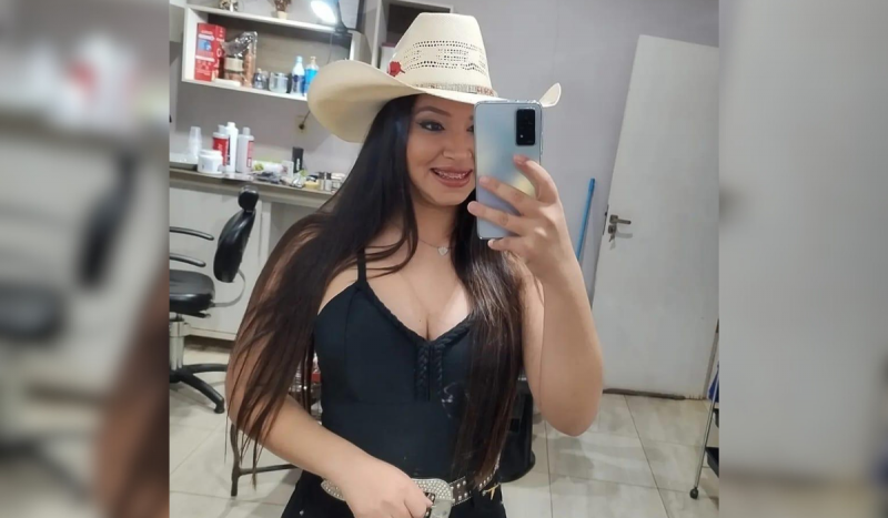 Mulher sorrindo tirando foto no espelho enquanto usa blusa decotada preta e chapéu de caubói na cor bege.