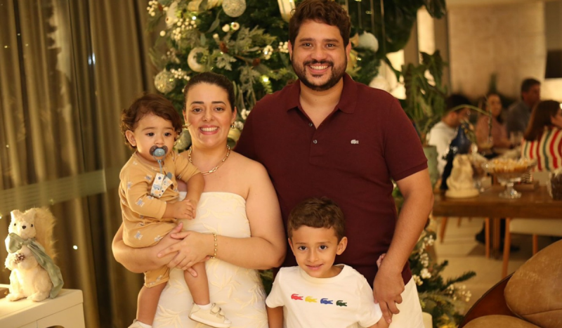 Mulher branca sorrindo ao lado de homem usando camisa polo vermelha. Ela carrega um bebê no colo e um outro menino sorri em pé na frente dela