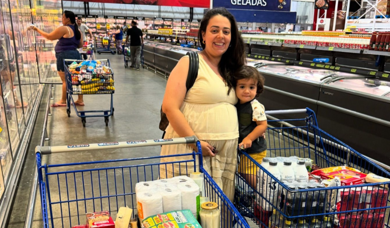 Influenciadora brasileira que teve embolia amniótica - foto mostra mulher branca, grávida sorrindo usando vestido bege longo ao lado de criança pequena em corredor de supermercado