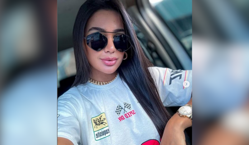 Foto de mulher branca usando camiseta branca. Ela tem cabelos lisos longos e pretos e está dentro de um carro usando óculos escuros