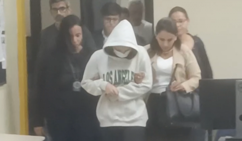 Mulher usando moletom branco cobrindo a cabeça sendo conduzida na delegacia após supostamente envenenar o namorado com um brigadeirão