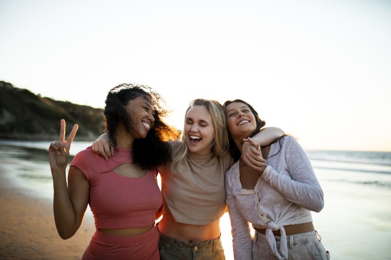 três amigas abraçadas e sorrindo em uma praia 