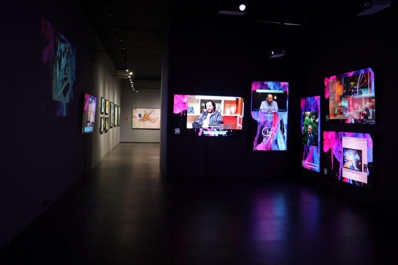 Foto de uma das salas do Museu da Imagem e do Som. A sala está todo escura e há cinco projeção coloridas em suas paredes. Ao fundo, é possível ver uma outra sala iluminada. Essa tem as paredes cinzas, onde estão fixados quadros coloridos. 