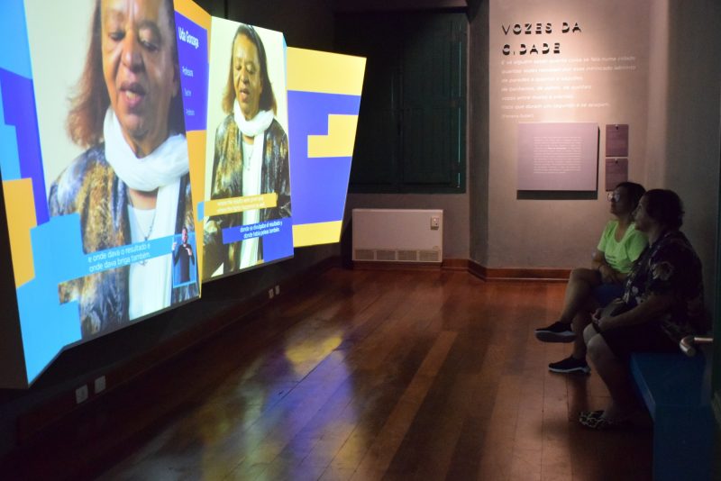 Foto de uma das salas do Museu de Florianópolis. No lado direito da foto, aparece um grande telão onde está sendo projetado um vídeo em que aparece uma senhora de pele preta e cabelos castanhos. Em frente ao telão, está um homem e uma mulher sentados em um banco, assistindo ao depoimento da senhora. Na parede do fundo está escrito "Vozes da cidade", seguida por um texto, que não aparece legível na foto. O chão da sala é de madeira. 