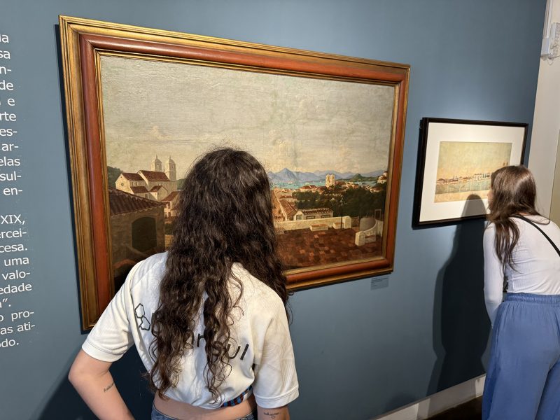 Foto de duas mulheres jovens observando dois quadros fixados em uma parede de cor cinza claro no Museu Victor Meirelles. Elas estão de costas para a foto. As duas tem pele branca e vestem blusas brancas. Uma delas tem cabelo longo, enrolado e preto, e a outra cabelo longo, liso e castanho.