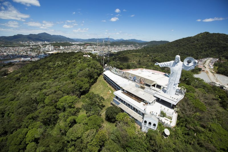 Foto do completo turístico Cristo Luz no alto do morro e cercado por vegetação. Ele aparece no lado centro-direito da foto. O complexo tem prédios brancos e a grande estátua do Cristo também é branca. Um de seus braços está esticado e no outro há um disco com holofote no centro. Ao fundo, aparece parte do centro urbano de Balneário Camboriú. 