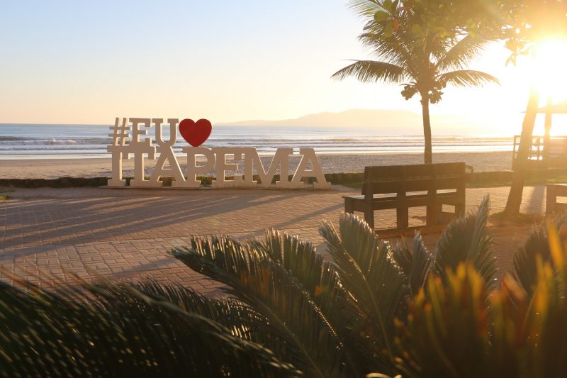 Foto de um calçadão na cidade de Itapema. No centro do calçadão, há um totem onde está escrito #Eu amo Itapema. A palavra ama é simbolizada por um coração vermelho. Ao lado, há um coqueiro e um banco de madeira. Atrás, o mar. Na parte inferior da foto, aparece algumas folhagens. O céu está azul e aparece a luz do sol no canto superior direito da foto. 