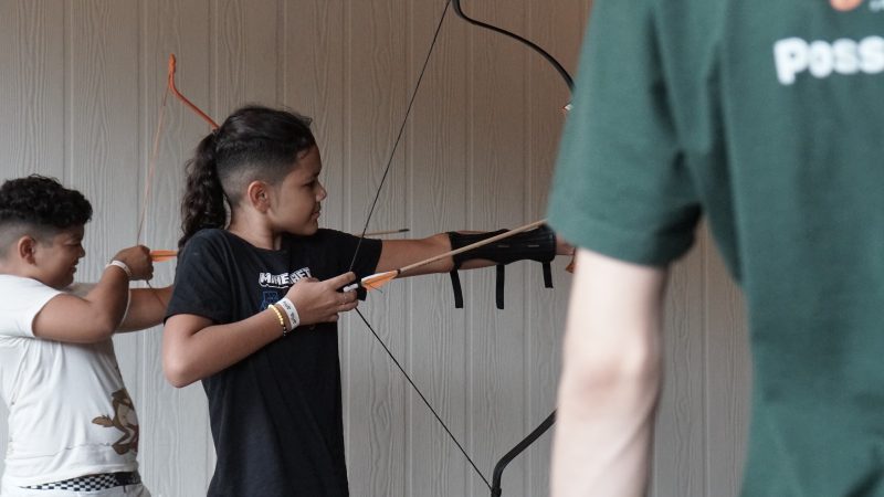 Foto de dois meninos de pele parda e cabelos pretos. Um deles veste uma blusa branca e o outro veste uma blusa preta. Eles estão segurando um arco e flecha e olham para frente, mirando o alvo, que não aparece na foto. 