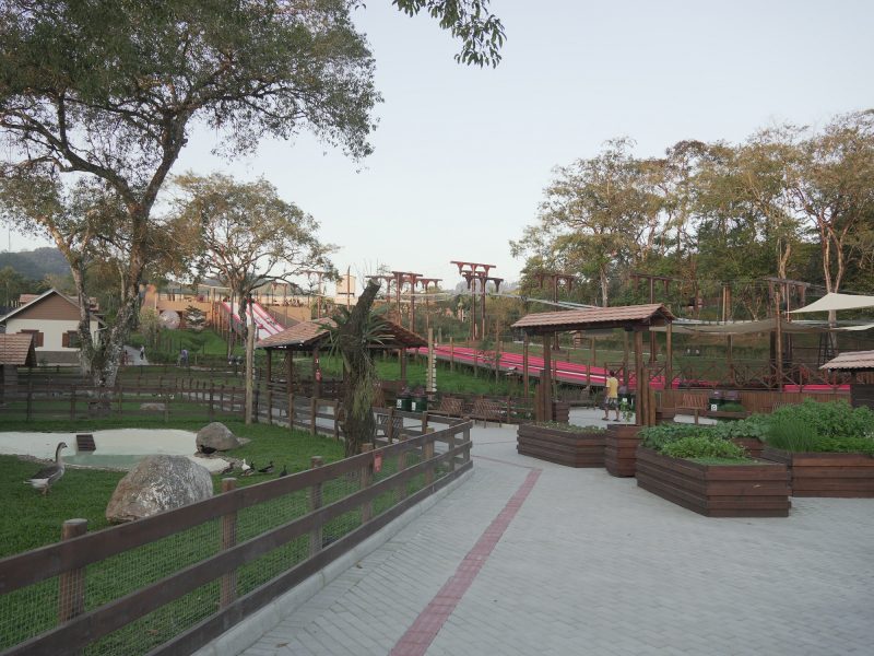 Foto geral do parque temático Spitz Pomer, em Pomerode. No lado esquerdo aparece uma área gramada, onde estão alguns patos, e cercada por uma estrutura de madeira. No lado direito, aparecem bancos de madeira e canteiros também de madeira onde estão algumas plantas. Ao fundo, aparecem árvores, parte da estrutura da tirolesa do parque e do escorregador gigante. 