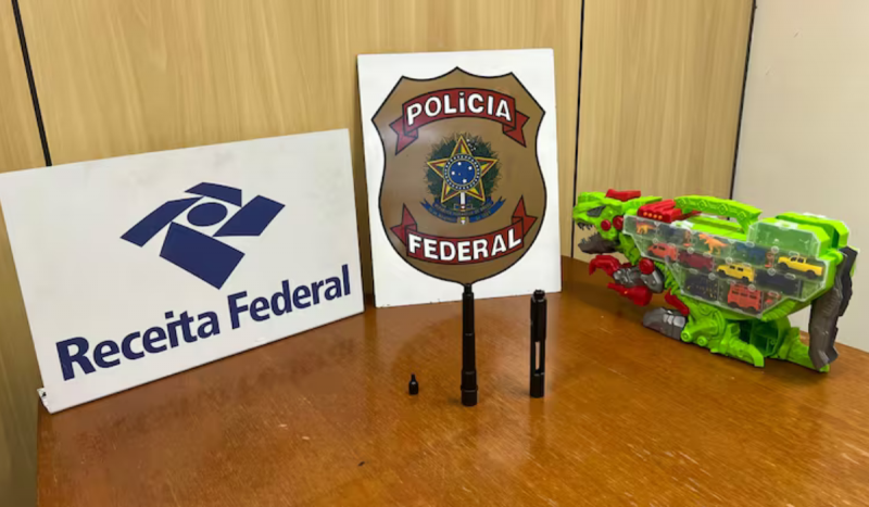 Peças de fuzil no centro de uma mesa de madeira marrom claro. Itens estão em frente à placas com as insígnias da PF e da Receita Federal, ao lado de brinquedo infantil da cor verde neon