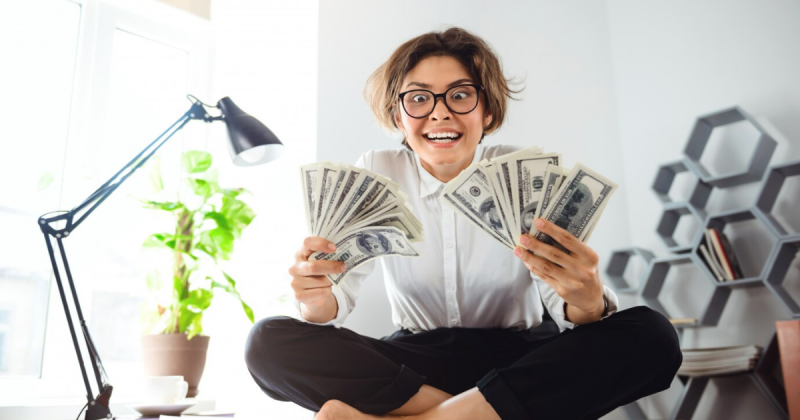 mulher de calça preta, blusa branca, óculos preto, cabelo amarrado meio desarrumado, sentada na posição de borboleta, segurando notas de dólares nas duas mãos, ela está com uma expressão de felicidade e surpresa