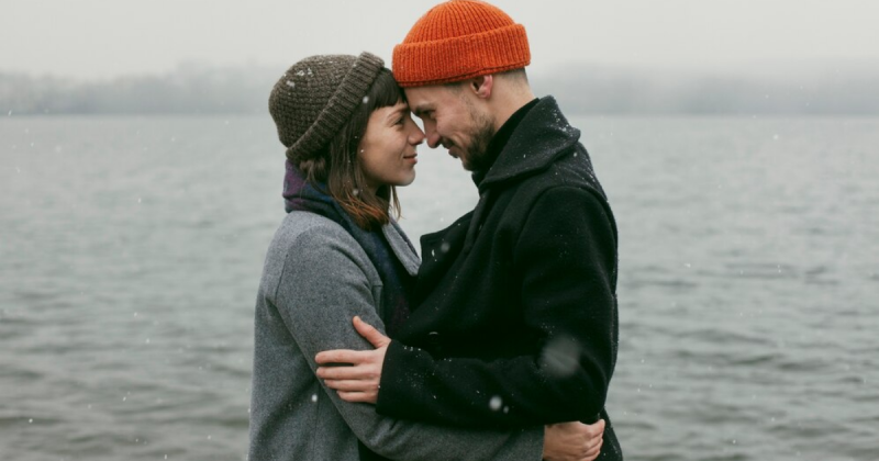 casal em frente a um lago abraçados e com a testa encostada uma na outra, os dois estão roupas de frio e touca 