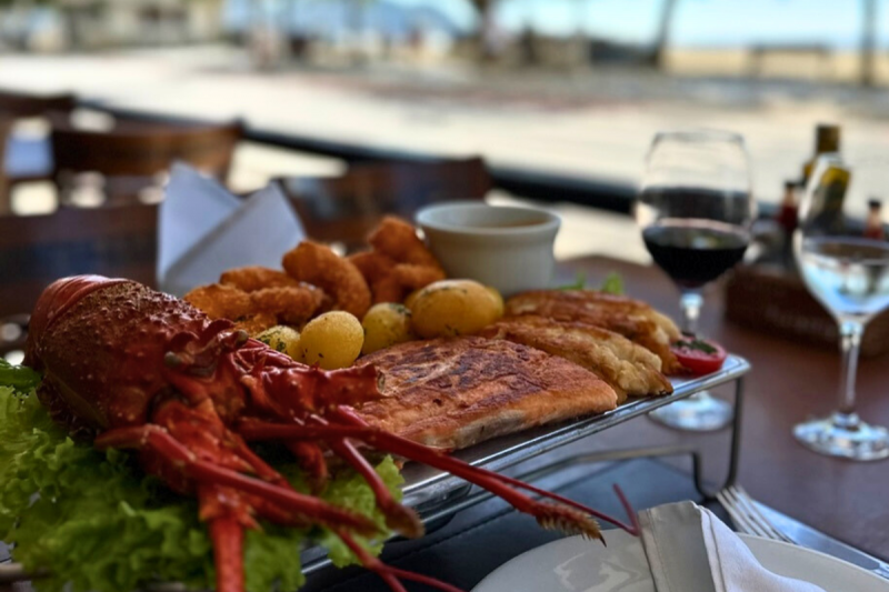 Foto da mesa do restaurante Casa da Lagosta, em Balneário Camboriú. Sobre a mesa está uma bandeja metálica sobre a qual há uma grande lagosta, filés de peixe e batatas. À frente aparece parte de um prato e duas taças. Ao fundo e desfocado aparece parte da areia da Praia Central. 