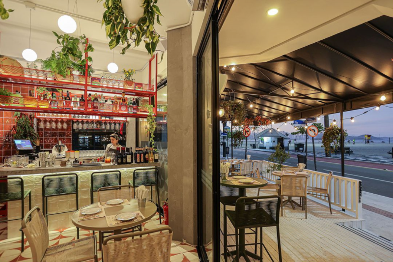 Foto do Restaurante Duladu Cozinha Italiana, em Balneário Camboriú. No lado esquerdo da foto aparece o ambiente interno do restaurante, com uma mesa pequena redonda e três cadeiras ao redor. Ao fundo, está um balcão com várias taças e bebidas. Uma moça jovem está atrás do balcão. No teto, estão três lustres pendentes de bolas brancas e algumas plantas penduradas. No lado direito da foto, aparece a varanda do restaurante, onde estão três pequenas mesas redondas. Ao fundo é possível ver parte da orla da Praia Central. 