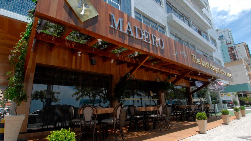 Foto da fachada do Madero Steak House, em Balneário Camboriú. A fachada do restaurante é toda em madeira e tem o nome escrito em letras metálicas. Ao lado do nome está escrito em inglês a seguinte frase: O melhor hambúrguer do mundo. Abaixo da fachada há uma varanda onde estão mesas feitas de madeira maciça e cadeiras. Grandes janelas de vidro escuro dividem a parte a externa e a interna do restaurante. 